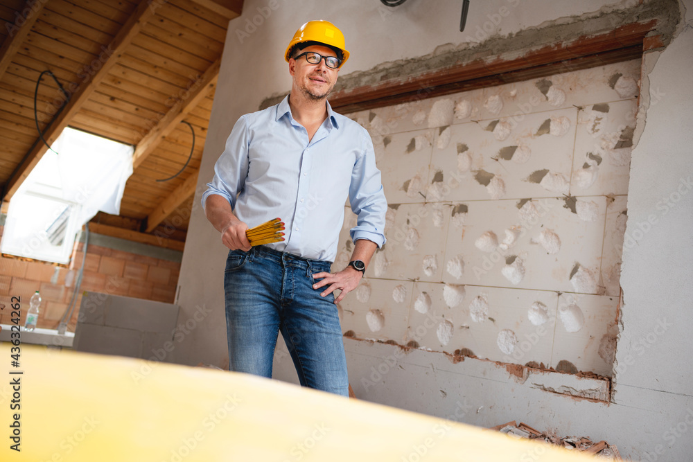 Architect with yellow safety helmet, blue shirt and jeans checks construction progress on building site in loft, attic with a yellow folding rule Architect with yellow safety helmet, blue shirt and jeans checks construction progress on building site in loft, attic with a yellow folding rule