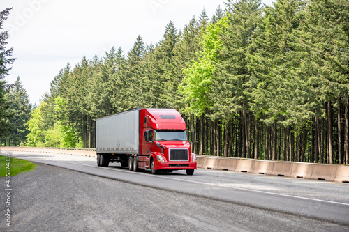 Popular red bonnet big rig semi truck with dry van semi trailer running on the divided highway road with green trees on the side