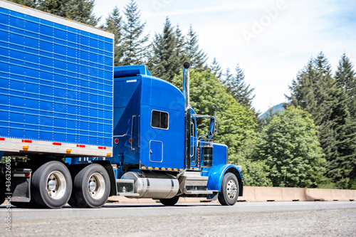 Stylish blue big rig classic bonnet semi truck with refrigerator semi trailer running on the divided highway road with trees on the side