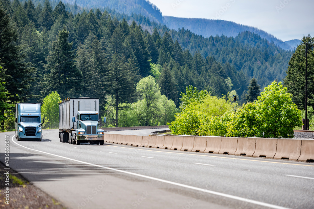 Two different big rig semi trucks with semi trailers running side by side on the winding multiline highway road with mountain and forest in Columbia Gorge
