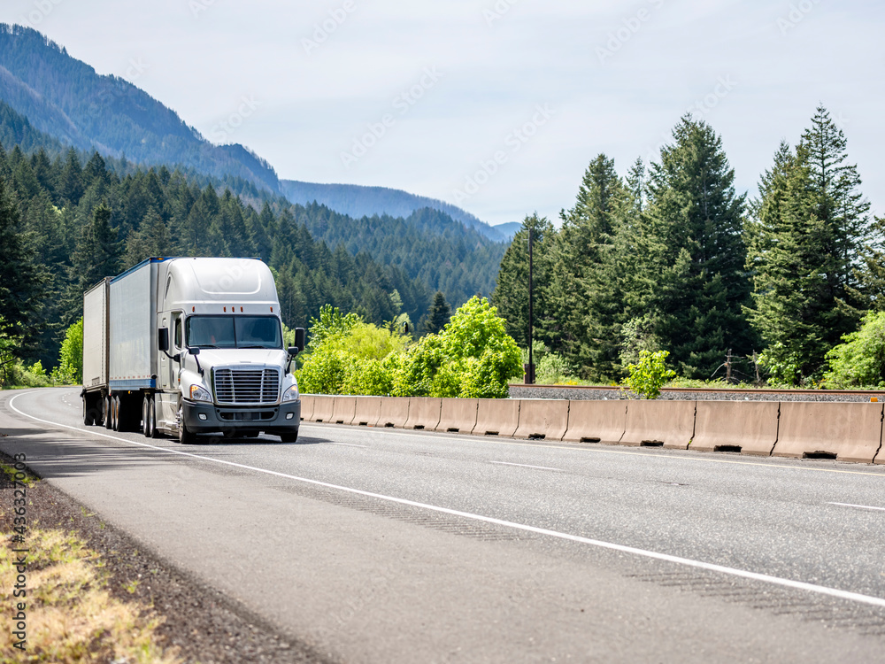 White bonnet big rig semi truck transporting cargo in two dry van semi trailers climbing on the winding mountain road with green forest around White bonnet big rig semi truck transporting cargo in two dry van semi trailers climbing on the winding mountain road with green forest around