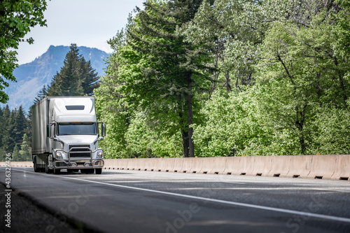 White powerful big rig semi truck with pipe grille guard transporting cargo in dry van semi trailer driving on the multiline highway green road