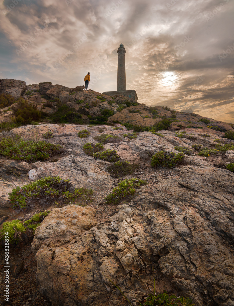 Adventurer standing looking at a lighthouse in a rocky land, Cabo de Palos, Murcia, Spain