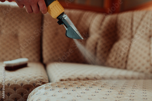 Close-up of housekeeper holding modern washing vacuum cleaner and cleaning dirty sofa with professionally detergent. Professional springclean at home concept photo