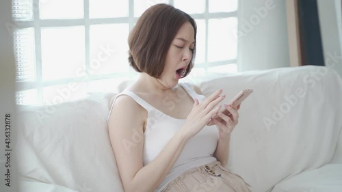lazy asian woman face yawning using mobile smart phone on sofa couch at home. female in white living room yawn on day time need rest. tired and exhausted from workload sleep fall asleep and nap.