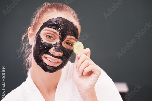 Smiling beautiful Caucasian woman in white bathrobe is holding cucumber slices and black facial mask on face for treatment skine care at spa photo