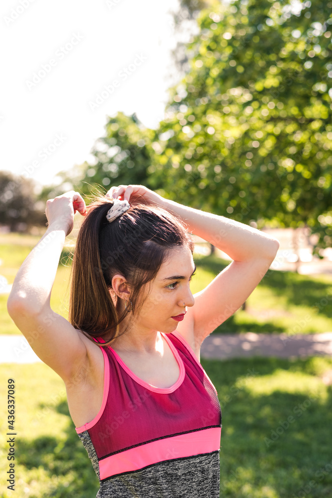 young caucasian woman collecting hair with a ponytail on a park. She is wearing ping and green sport clothes
