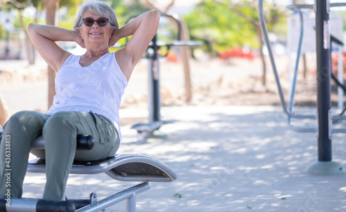Attractive senior woman in sport activity in public park, sitting on fitness equipment doing exercises, smiling