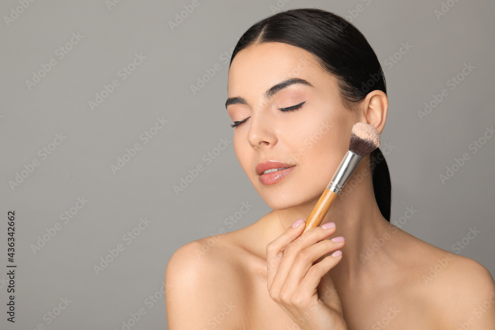 Fototapeta premium Beautiful young woman applying face powder with brush on grey background. Space for text