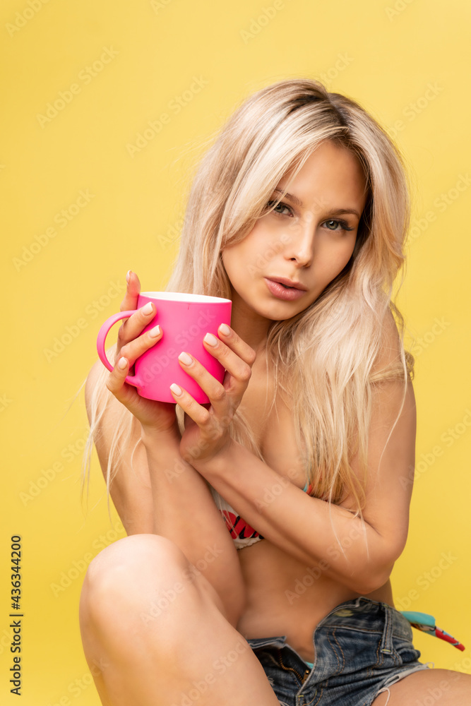 A girl sitting on the floor with her feet cross-legged keeps her head on a beautiful yellow background with a cup in hand yellow or pink and red playful swimsuit sexy, figure hair looking person, hot A girl sitting on the floor with her feet cross-legged keeps her head on a beautiful yellow background with a cup in hand yellow or pink and red playful swimsuit sexy, figure hair looking person, hot