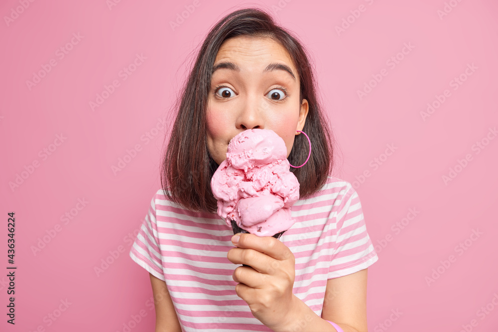 Photo of surprised Asian woman licks delicious ice cream has sweet tooth eats high calorie food dressed in casual striped t shirt during summer isolated on pink background. Treat yourself with dessert - obrazy, fototapety, plakaty 