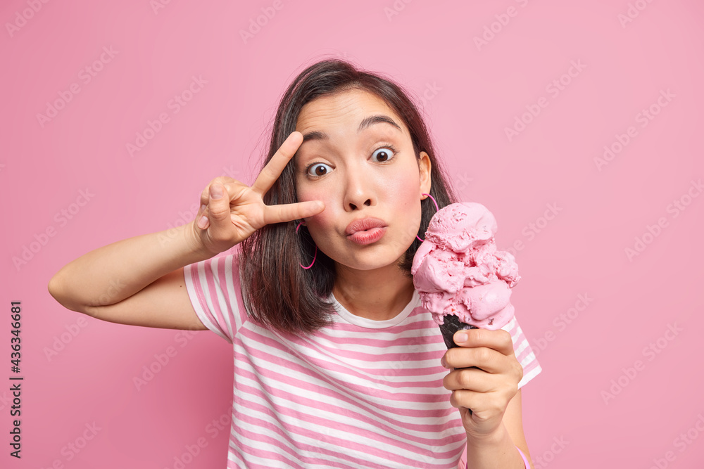 Surprised lovely Asian woman keeps lips folded makes peace gesture over eye dressed in striped t shirt holds tasty ice cream for sweet tooth isolated over pink background. People lifestyle concept