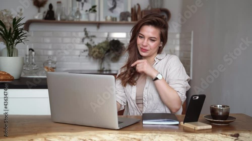 Young beautiful laughing woman talking on a video call with friends and family sitting at the table in the kitchen.