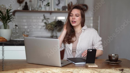 Smiling young blonde woman in beige linen clothes with her laptop computer,uses a mobile phone, sitting at a table indoors, talking on a video call.Home schooling in college.Freelancer works from home