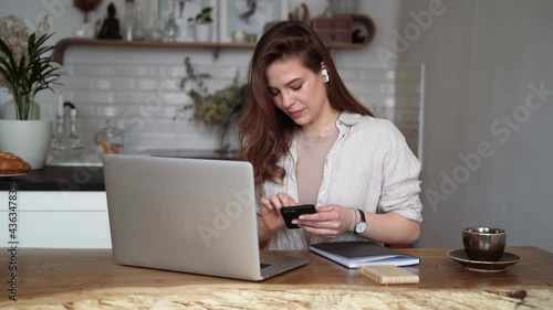 Smiling young blonde woman in beige linen clothes with her laptop computer,uses a mobile phone, sitting at a table indoors, talking on a video call.Home schooling in college.Freelancer works from home