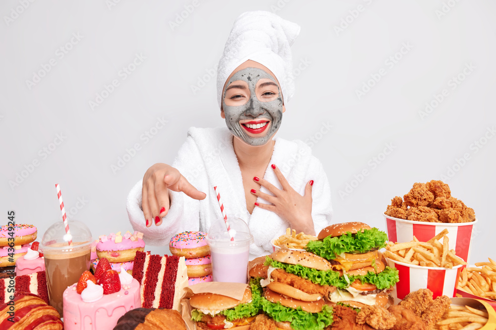 Positive woman applies clay mask for reducing fine lines smiles broadly has good mood dressed in bathrobe poses against different fast food snacks isolated over white background. Harmful nutrition Positive woman applies clay mask for reducing fine lines smiles broadly has good mood dressed in bathrobe poses against different fast food snacks isolated over white background. Harmful nutrition