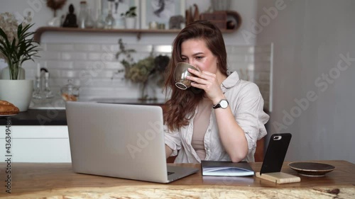 Smiling young blonde woman in beige linen clothes with her laptop computer, drinking coffee, sitting at a table indoors, talking on a video call. Homeschooling. Freelancer works from home