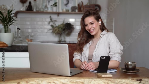 Smiling young blonde woman in beige linen clothes with her laptop computer, drinking coffee, sitting at a table indoors, talking on a video call. Homeschooling. Freelancer works from home