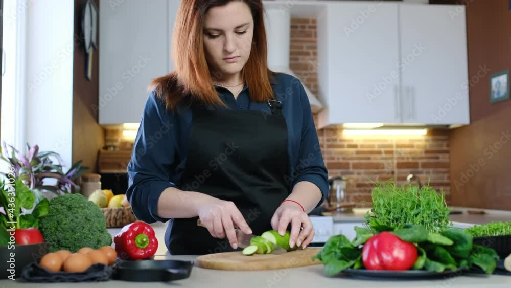 concept of food experiences at home cooking and at-home dining. a redheaded woman in a black apron cuts chukini for shakshuka in her kitchen concept of food experiences at home cooking and at-home dining. a redheaded woman in a black apron cuts chukini for shakshuka in her kitchen