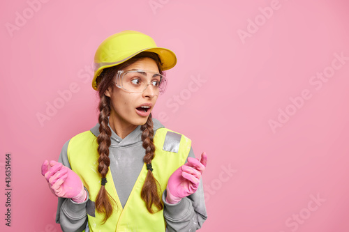 Repair and construction concept. Puzzled surprised woman spreads hands loks away notices something amazing wears protective hardhat transparent safety glasses and uniform isolated on pink wall photo