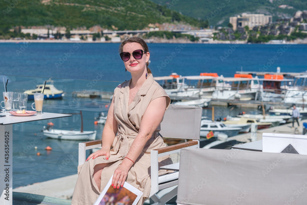 Young pretty European woman in sunglasses with brown hair pulled back is sitting at in a terrace of a summer cafe with sea and marina view and holds a magazine, laughing