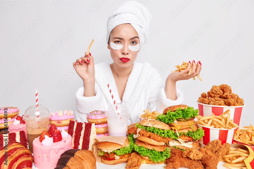 Pensive Asian woman focused at camera eats fries has red lips applies patches under eyes surrounded by junk food keeps to unhealthy nutrition isolated over white background doesnt care about diet Pensive Asian woman focused at camera eats fries has red lips applies patches under eyes surrounded by junk food keeps to unhealthy nutrition isolated over white background doesnt care about diet