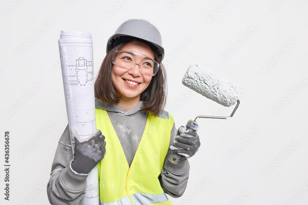Horizontal shot of positive female construction worker smiles positively  holds paint roller and blueprint being in good mood ready to start working  isolated over white background. Industrial engineer Stock Photo | Adobe
