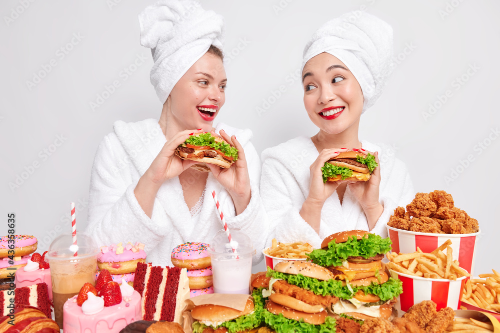 Junk food consuming. Positive two women best friends eat delicious hamburgers stand closely to each other surrounded by different tasty high calorie snacks isolated over white background. Big portions Junk food consuming. Positive two women best friends eat delicious hamburgers stand closely to each other surrounded by different tasty high calorie snacks isolated over white background. Big portions