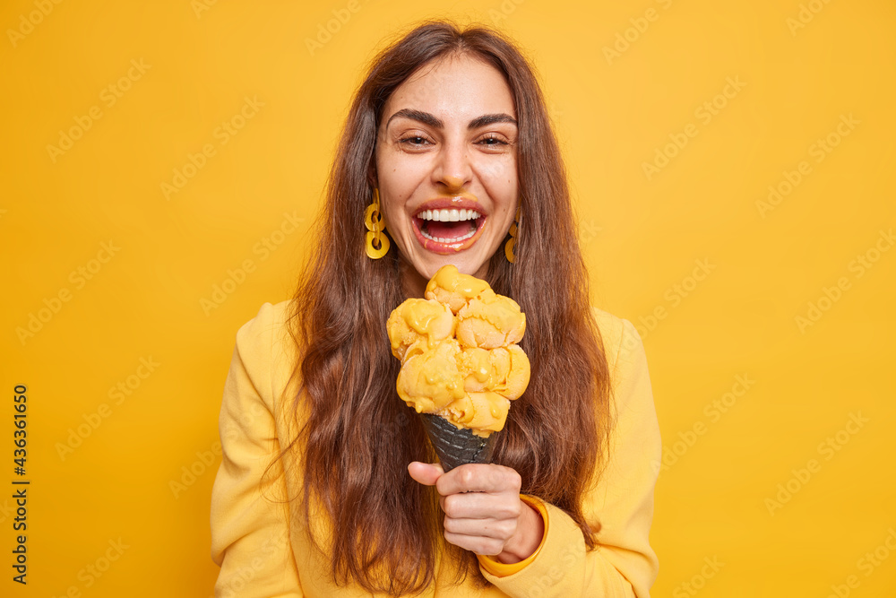 Φωτογραφία Cheerful young brunette European woman smiles gladfully eats appetizing ice cream perfect for summer heat smiles broadly dressed in jacket isolated over yellow background