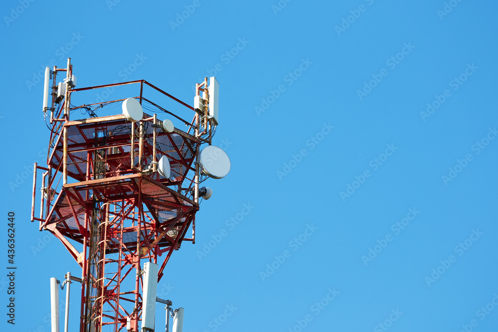 A telephone metal tower of red and white radio communication hung with equipment and satellite dishes against the blue sky. Modern technology and communication communications. Copy space. A telephone metal tower of red and white radio communication hung with equipment and satellite dishes against the blue sky. Modern technology and communication communications. Copy space.
