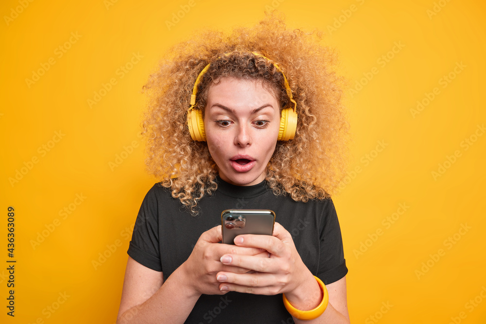 Studio shot of impressed curly haired woman focused at smartphone display feels very surprised reads text message dressed in casual black t shirt isolated over yellow background. Human reactions Studio shot of impressed curly haired woman focused at smartphone display feels very surprised reads text message dressed in casual black t shirt isolated over yellow background. Human reactions