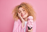 Portrait of lovely young European woman looks with delight at camera keeps hand on cheek smiles gently holds tasty big ice cream in waffle dressed in stylish jacket isolated over pink background.