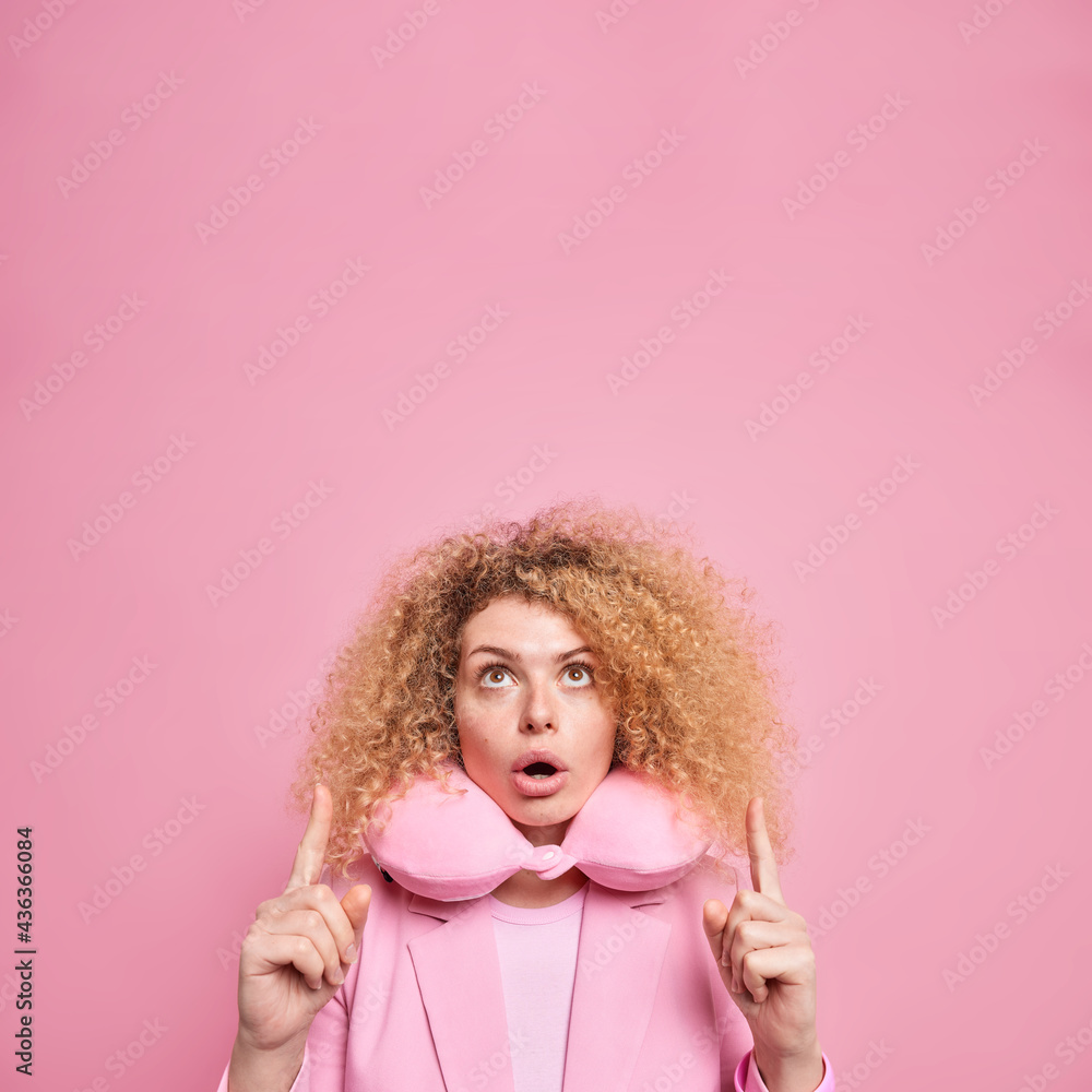 Surprised woman with curly bushy hair feels impressed points above on copy space wears travel pillow to feel comfort for neck dressed in elegant clothes isolated over pink background. Your promo here Surprised woman with curly bushy hair feels impressed points above on copy space wears travel pillow to feel comfort for neck dressed in elegant clothes isolated over pink background. Your promo here