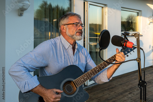 Senior man imposing musician plays the guitar and sings into microphone. Elegant elderly male 60 bearded performs outdoors in summer. Attractive handsome pensioner man activity creativity.