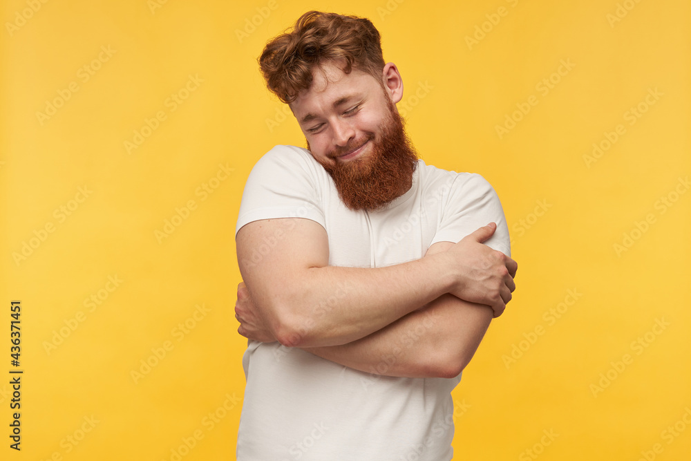 indoor portrait of young bearded guy, wears blank t-shirt, keeps his eyes closed, hug himself and smiling. isolated over yellow background indoor portrait of young bearded guy, wears blank t-shirt, keeps his eyes closed, hug himself and smiling. isolated over yellow background