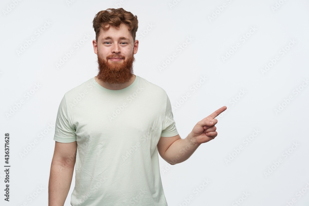 indoor shot of young bearded redhead guy, wears blank t-shirt, pointing with a finger at right side copy space, smiling and has amazed facial expression. isolated over white background. indoor shot of young bearded redhead guy, wears blank t-shirt, pointing with a finger at right side copy space, smiling and has amazed facial expression. isolated over white background.