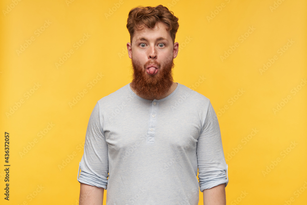 indoor shot of young funny bearded male with a stylish haircut, keeps his eyes opened and show his tongue. isolated over yellow background. indoor shot of young funny bearded male with a stylish haircut, keeps his eyes opened and show his tongue. isolated over yellow background.