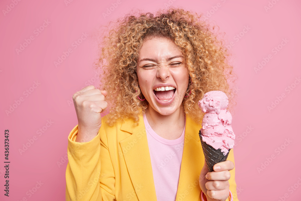 Overjoyed curly haired woman makes triumph gesture clenches fist and exclaims from joy holds big appetizing ice cream dressed in yellow jacket isolated over pink background. People and happiness Overjoyed curly haired woman makes triumph gesture clenches fist and exclaims from joy holds big appetizing ice cream dressed in yellow jacket isolated over pink background. People and happiness