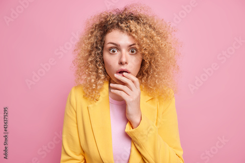 Portrait of surprised young woman with curly bushy hair stands ambushed stares speechless with startled expression hears shocking unbelievable newswears formal yellow jacket isolated on pink wall