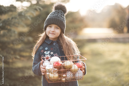 Cute funny child girl 4-5 year old holding Christmas decorations in basket wear winter coat and knitted hat in park outdoors. WInter holiay season. Looking at camera. Happy kid with xmas decor photo