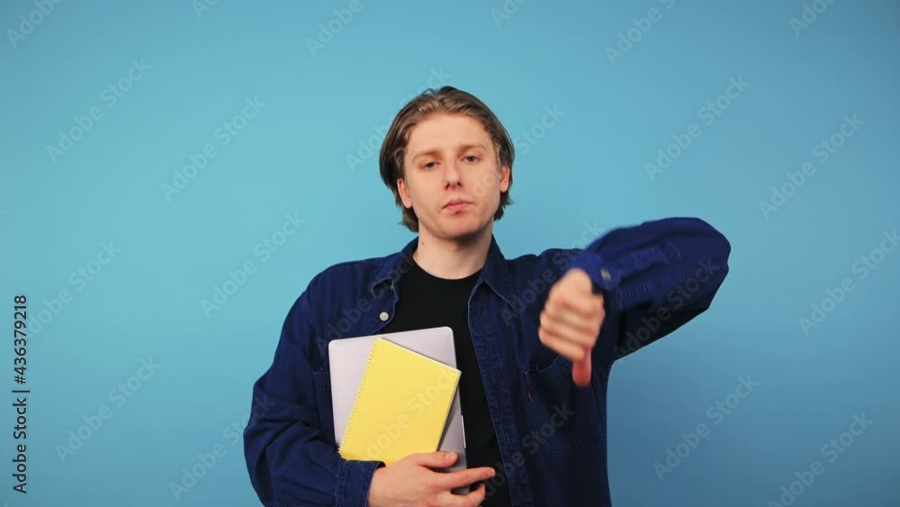 Dissatisfied student man in a shirt stands on a blue background and shows a dislike gesture with his fingers with a displeased face looking at the camera. Dissatisfied student man in a shirt stands on a blue background and shows a dislike gesture with his fingers with a displeased face looking at the camera.