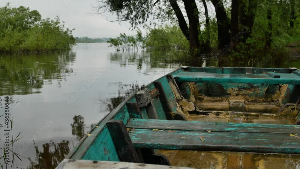 4K summer video background. An old wooden small fishing boat moored by the river on a rainy summer or spring day against the backdrop of a large river and green trees. 4K summer video background. An old wooden small fishing boat moored by the river on a rainy summer or spring day against the backdrop of a large river and green trees.