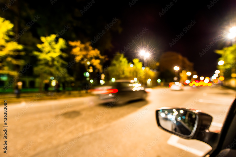Night city lights reflected in the mirror of the car, night avenue in the light of lanterns and passing  car. Wide-angle view and defocused image, from the car window