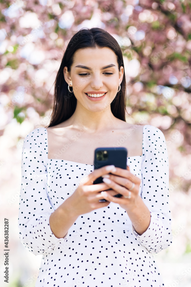 Happy hipster woman typing by mobile phone outdoors. Cheerful girl with smartphone in park on a background of sakura trees. Smiling lady holding cellphone in hands outside Happy hipster woman typing by mobile phone outdoors. Cheerful girl with smartphone in park on a background of sakura trees. Smiling lady holding cellphone in hands outside