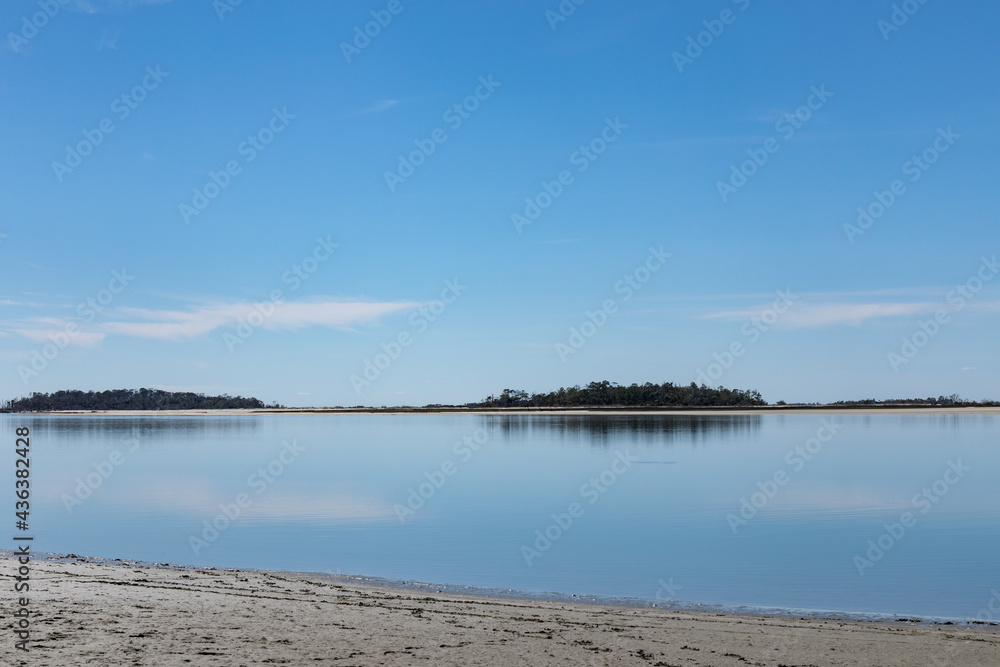Sandy beach by still water, distant islands, blue sky reflected in the water, horizontal aspect