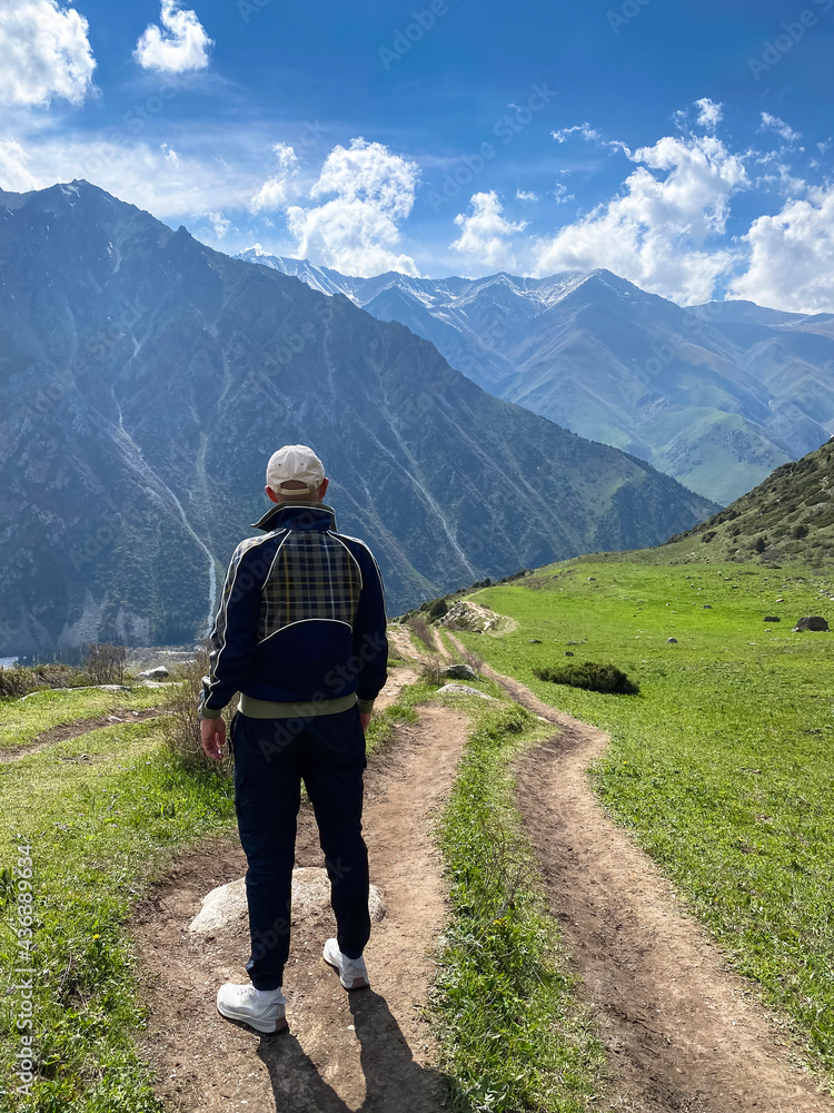 A beautiful mountain landscape and a path in a meadow among green grass. A tourist, a man in sports clothes is walking along a mountain path