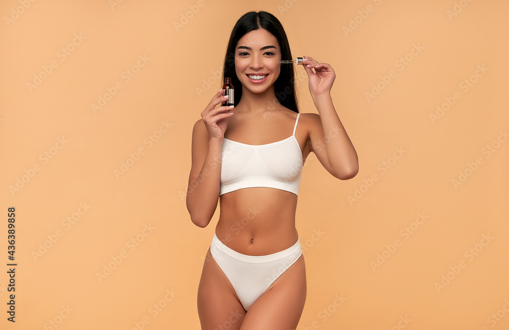 Fototapeta premium Young Asian woman with clean radiant skin in white lingerie uses a moisturizing serum on a beige background. Spa care, facial skin care, beauty cosmetology.