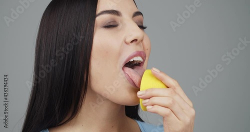 Beautiful young woman licking half of soar lemon on grey background photo