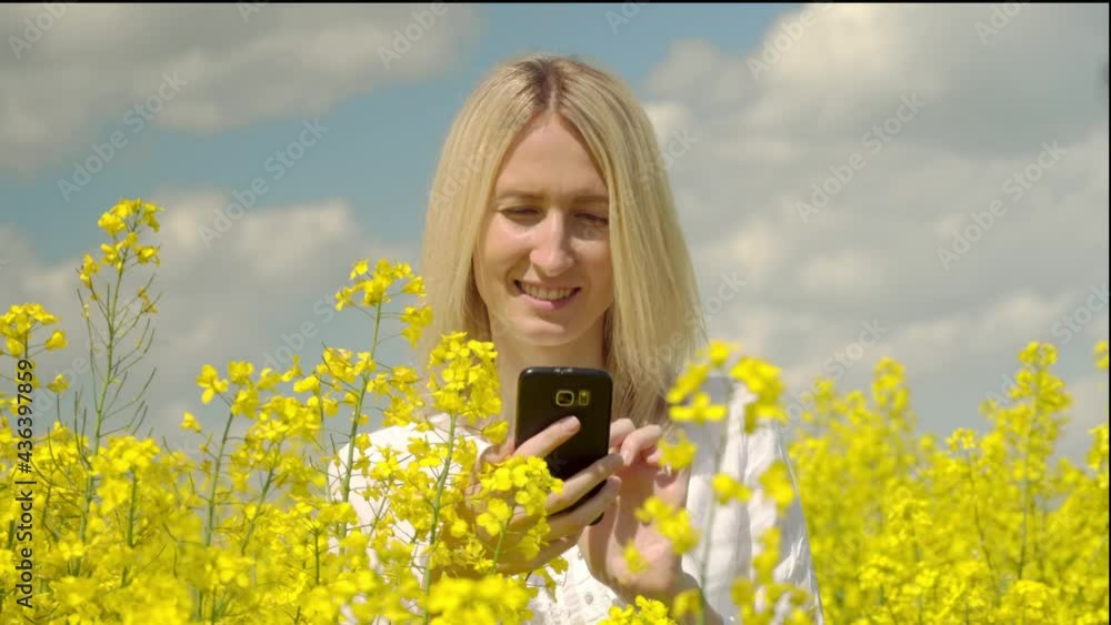 A young beautiful blonde takes photos of a flowering rapeseed in a field in the open air. She takes photos on her smartphone in the countryside. A happy woman. A young beautiful blonde takes photos of a flowering rapeseed in a field in the open air. She takes photos on her smartphone in the countryside. A happy woman.