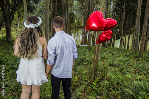 couple holding hands and back in their pre wedding in nature in a forest holding red heart balloons.